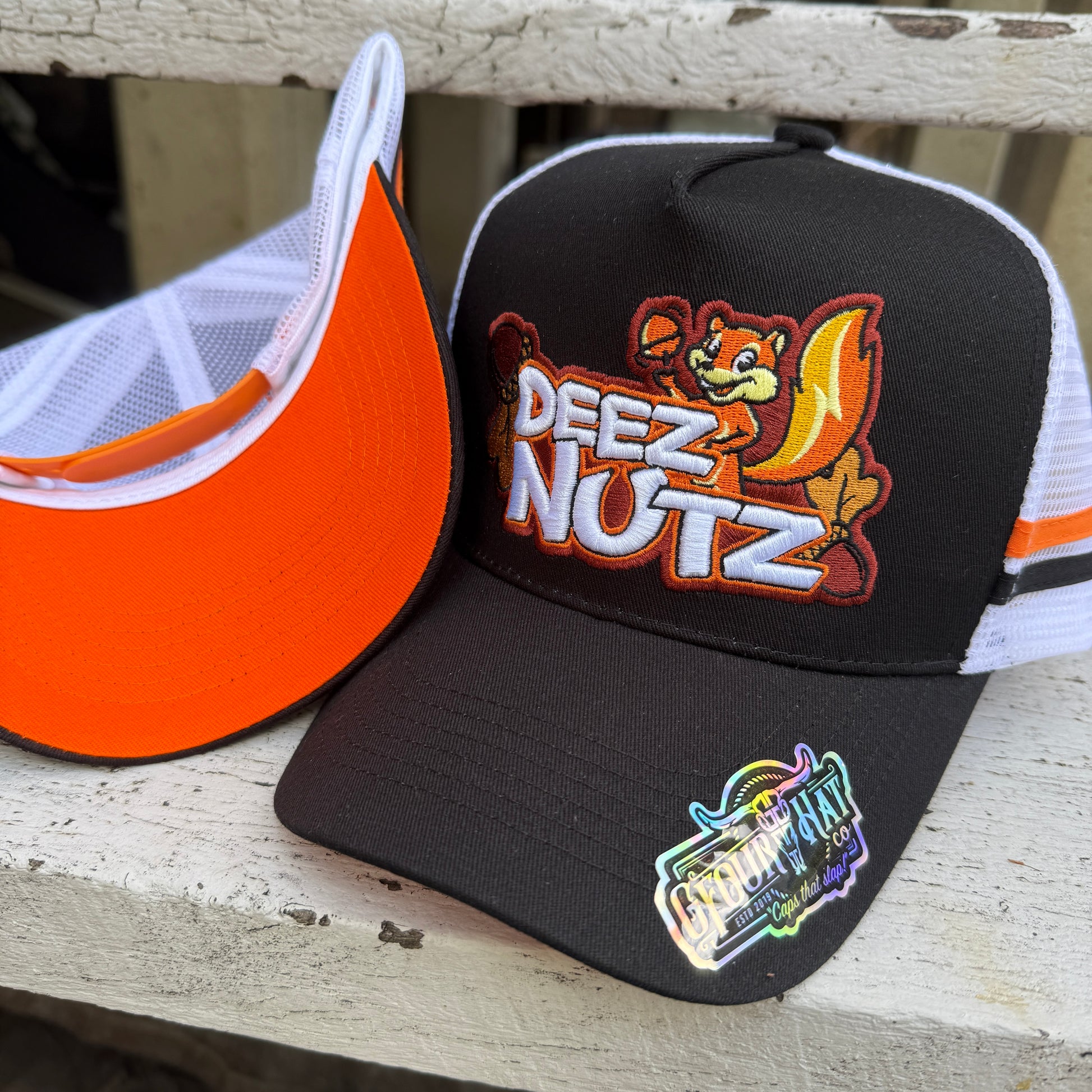 A black 'Deez Nutz' trucker cap with an orange visor and white mesh back, featuring an animated squirrel design and text logo.